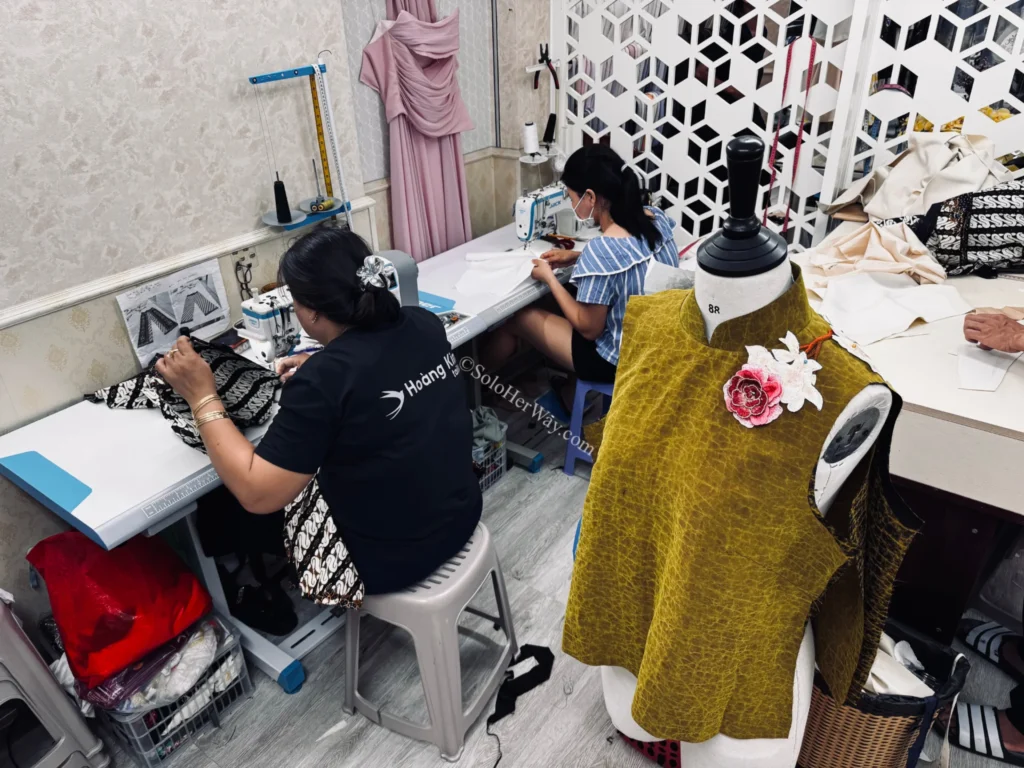 Getting Clothes Made in Hoi An & Ho Chi Minh City: What I Wish I’d Known 4 Tailors sewing custom garments inside a busy Vietnam tailoring shop with dress forms, fabrics, and sewing machines in active use.