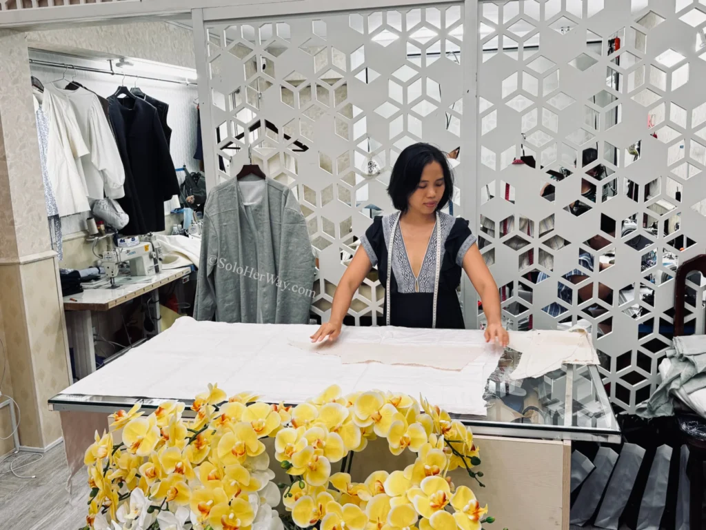 Getting Clothes Made in Hoi An & Ho Chi Minh City: What I Wish I’d Known 3 Tailor arranging fabric patterns on a cutting table inside a custom clothing workshop with garments and sewing equipment visible in the background.